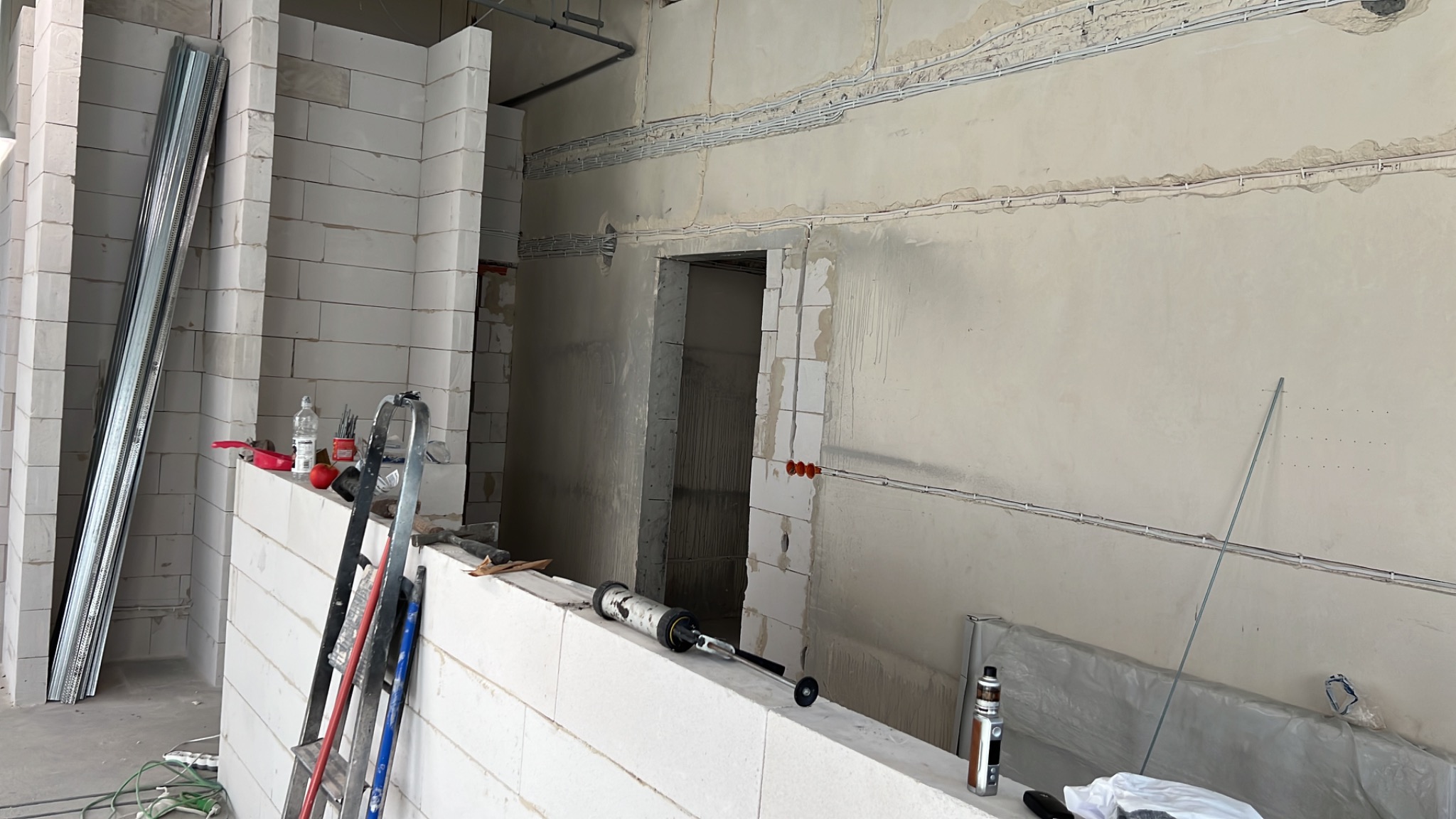 Wnętrze w trakcie budowy z widocznymi przewodami elektrycznymi na ścianach, narzędziami i materiałami budowlanymi, w tym profile metalowe i bloczki z betonu komórkowego.