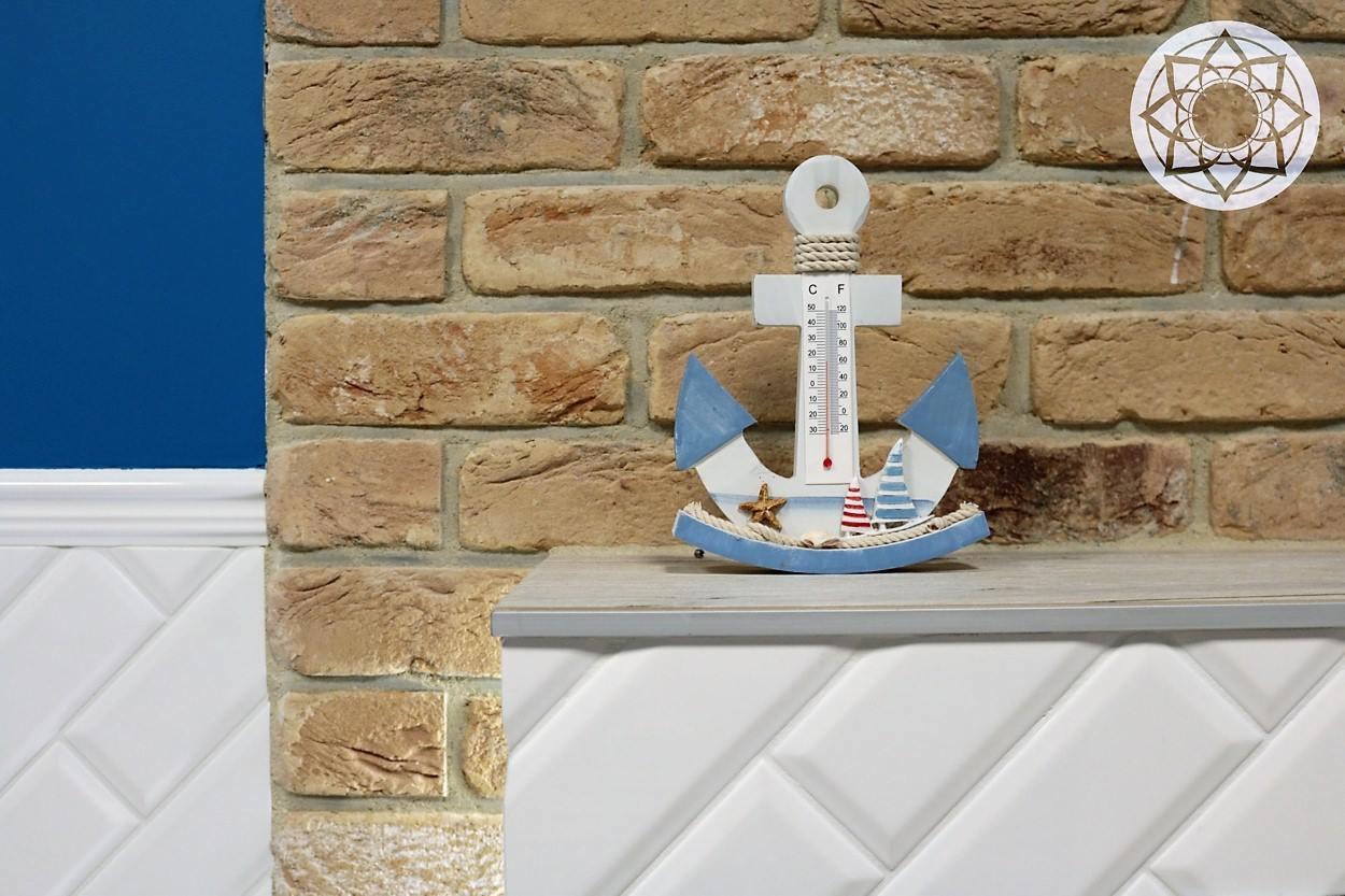 Dekoracyjny termometr w kształcie kotwicy na tle ściany z cegły klinkierowej i białych, ukośnych płytek ceramicznych.