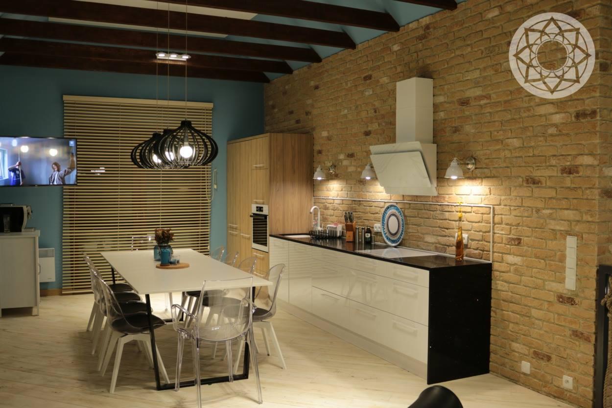 Nowoczesna kuchnia z jadalnią w kamienicy: białe meble, ceglana ściana z dekoracyjnym ornamentem, designerskie lampy wiszące i przezroczyste krzesła.