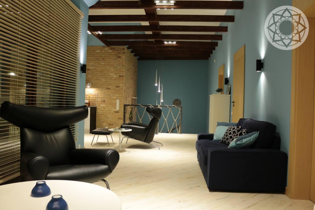 Nowoczesny salon z drewnianymi belkami sufitowymi, ceglaną ścianą, designerskimi fotelami i kanapą w odcieniach granatu i błękitu, oświetlony punktowo z żaluzjami w oknach.