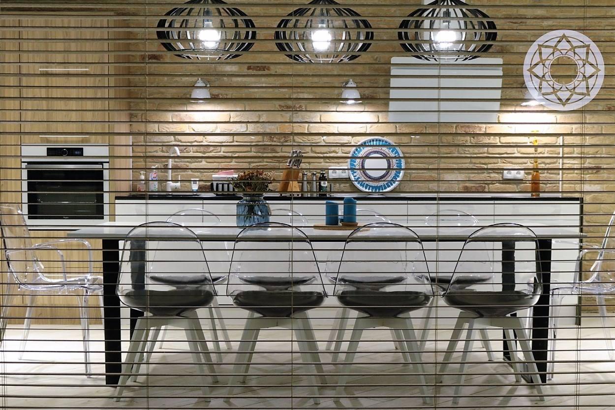 Nowoczesna kuchnia z ceglaną ścianą, oświetlona trzema designerskimi lampami wiszącymi i dodatkowymi kinkietami, widoczna przez drewniane żaluzje.