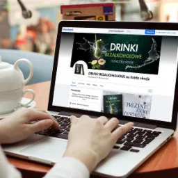 Osoba pisze na laptopie, na ekranie strona profilowa na Facebooku marki bezalkoholowych drinków z zielonym koktajlem na banerze, obok dzbanek z herbatą i deser.