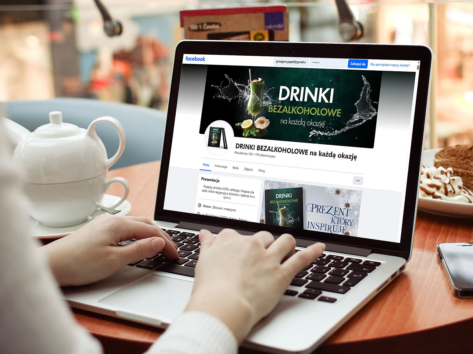 Osoba pisze na laptopie, na ekranie strona profilowa na Facebooku marki bezalkoholowych drinków z zielonym koktajlem na banerze, obok dzbanek z herbatą i deser.