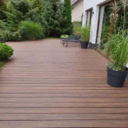 Brązowy taras z desek kompozytowych, z widocznymi roślinami w donicach oraz leżakami, przylegający do białego budynku z dużymi oknami.