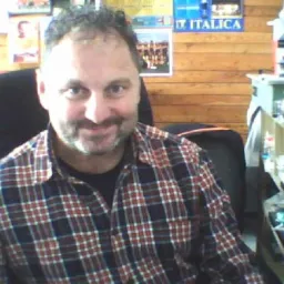 Portret uśmiechniętego mężczyzny w kraciastej koszuli na tle drewnianej ściany z plakatami i półką. Domowe biuro.