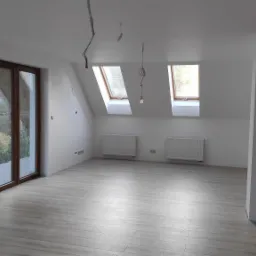 Wnętrze poddasza po remoncie: świeżo pomalowane ściany na biało, drewniane okna dachowe, grzejniki, drewnopodobna podłoga, kolumna z cegły klinkierowej.