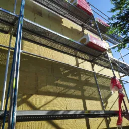 Ściana budynku w trakcie ocieplania wełną mineralną, widoczne rusztowanie z materiałami izolacyjnymi Rockwool na górnych poziomach, niebieska lina zabezpieczająca i częściowo zamontowana izolacja.