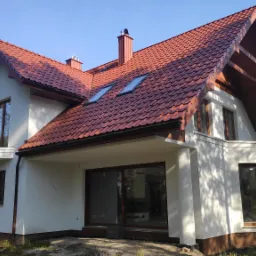 Nowoczesny dom jednorodzinny z białą elewacją, czerwoną dachówką, drewnianą podbitką i oknami w brązowych ramach, widok z zewnątrz w słoneczny dzień.