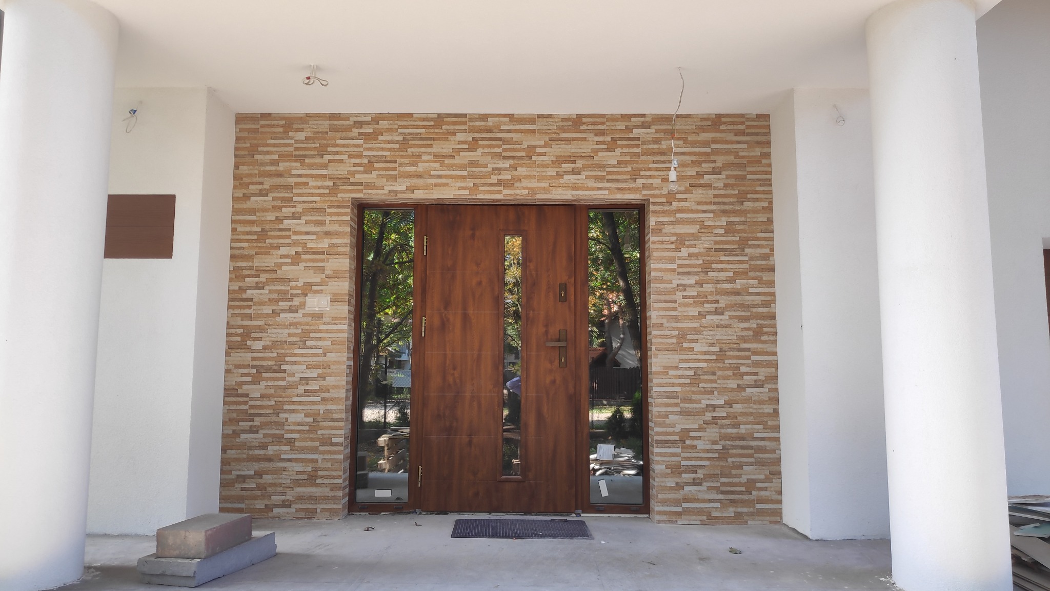 Wejście do budynku z drewnianymi drzwiami, otoczone elewacją z cegiełek w odcieniach beżu i brązu, wsparte na dwóch białych, okrągłych kolumnach. Widoczne niedokończone prace wykończeniowe na podłożu.