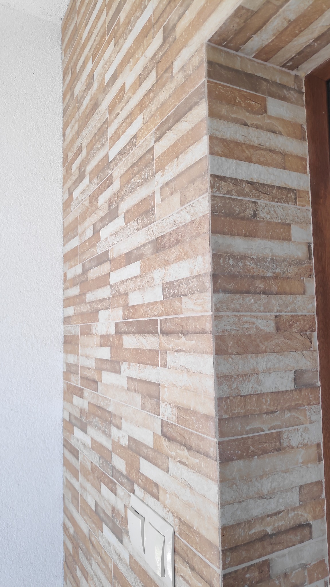 Ściana wykończona podłużnymi, beżowo-brązowymi płytkami imitującymi kamień, z widocznym narożnikiem i białym włącznikiem światła, sąsiadująca z białą, teksturowaną ścianą.