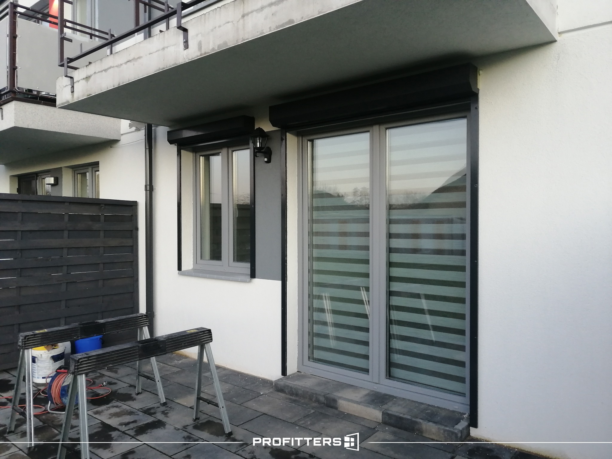 Szary budynek z zamontowanymi roletami dzień i noc oraz roletami zewnętrznymi, widoczne rusztowania malarskie i akcesoria.