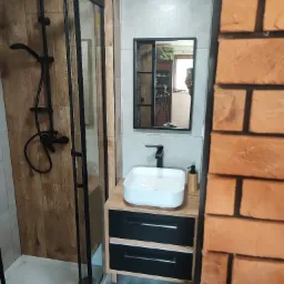 Nowoczesna łazienka z kabiną prysznicową typu walk-in z czarną armaturą, umywalką nablatową na szafce z drewnianym blatem i czarnymi frontami oraz lustrem w czarnej ramie, widok z perspektywy...