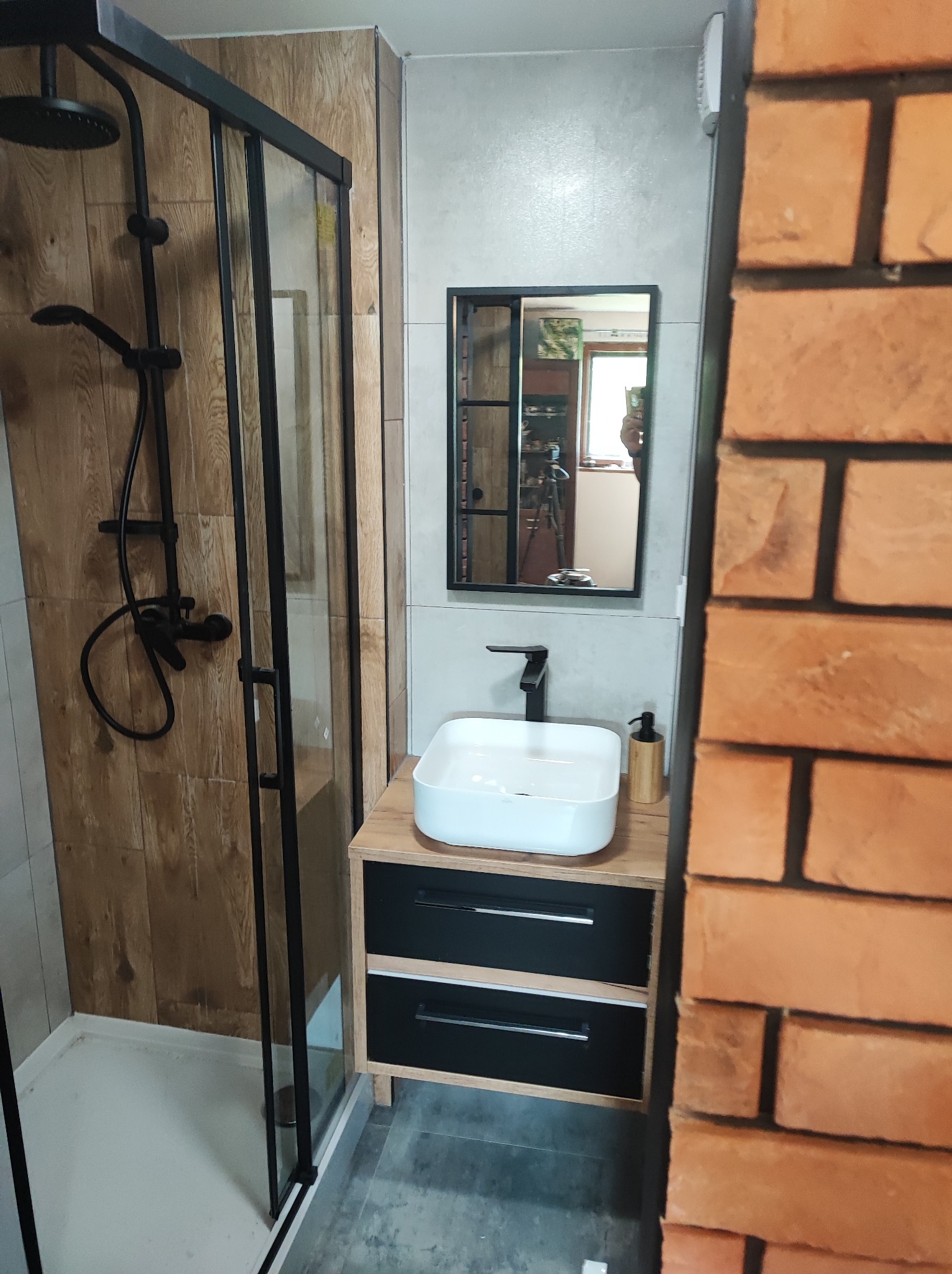 Nowoczesna łazienka z kabiną prysznicową typu walk-in z czarną armaturą, umywalką nablatową na szafce z drewnianym blatem i czarnymi frontami oraz lustrem w czarnej ramie, widok z perspektywy...