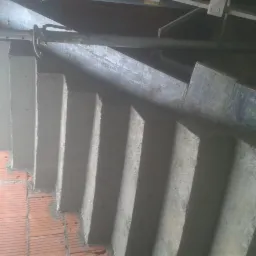 Surowe betonowe schody w trakcie budowy, widoczne cegły i stalowe podpory, oświetlenie budowlane.