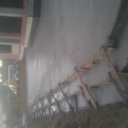 Świeżo wylana, mokra posadzka betonowa wewnątrz budynku w trakcie budowy, widoczne drewniane szalunki i fragmenty zbrojenia, w tle fragment muru i okna.