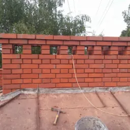 Ceglany komin z otworami wentylacyjnymi na miedzianym dachu, widoczne narzędzia i wiadro, drzewa w tle.