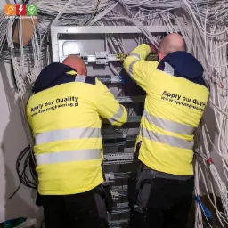 Dwóch elektryków w żółtych kurtkach firmowych pracuje nad podłączeniem kabli w skrzynce rozdzielczej w pomieszczeniu z widocznymi przewodami.