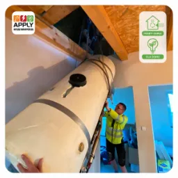 Montaż zbiornika buforowego pompy ciepła w przestrzeni poddasza, pracownik w odblaskowej kurtce przytrzymuje urządzenie zawieszone na linach, widoczne elementy konstrukcji dachu i logo firmy Apply...