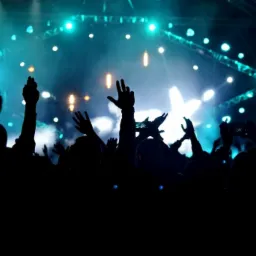 Tłum ludzi z podniesionymi rękami w ciemności, oświetlony niebieskimi i białymi światłami sceny podczas koncertu.