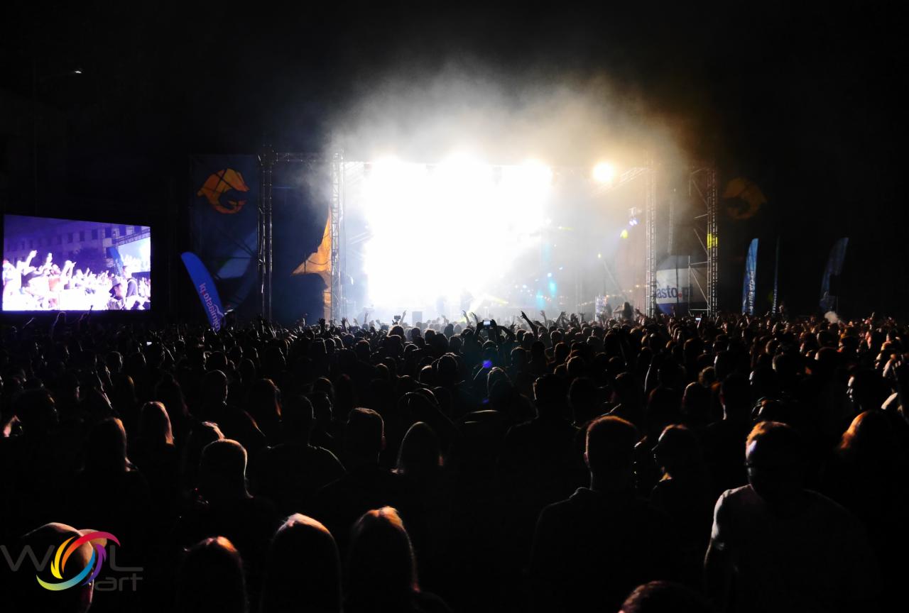 Tłum ludzi na koncercie z podniesionymi rękami, scena z oświetleniem i telebimem w tle.