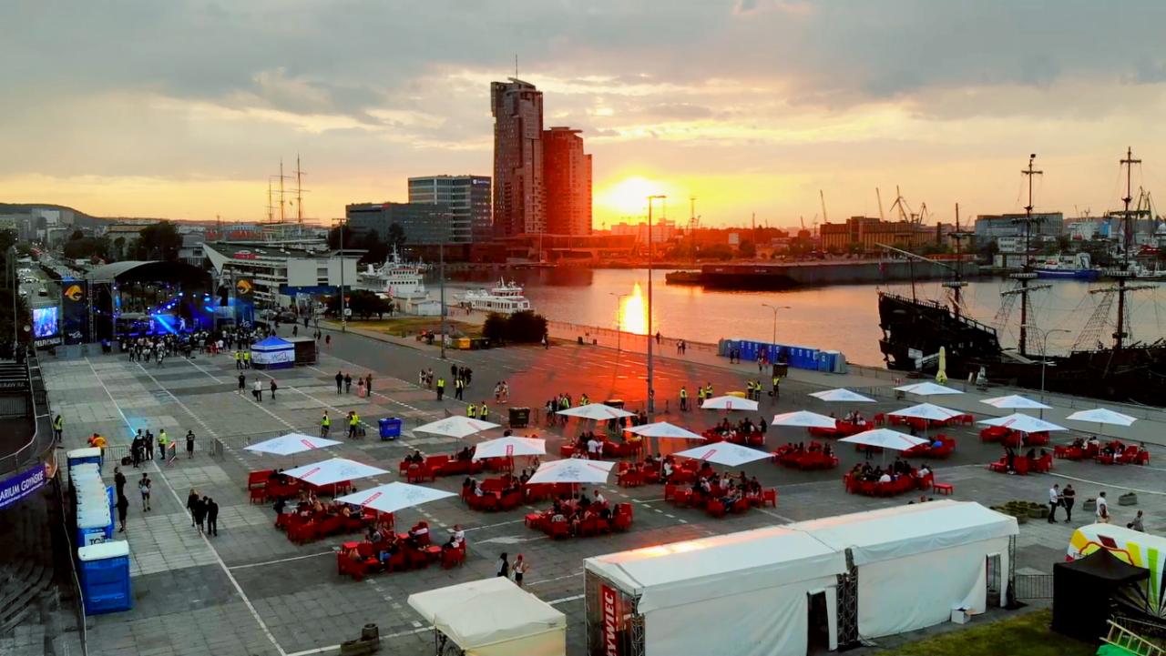 Widok z góry na plac miejski w Gdyni podczas wydarzenia plenerowego o zachodzie słońca, z licznymi stolikami pod parasolami dla uczestników, sceną koncertową w tle i żaglowcem przy nabrzeżu.