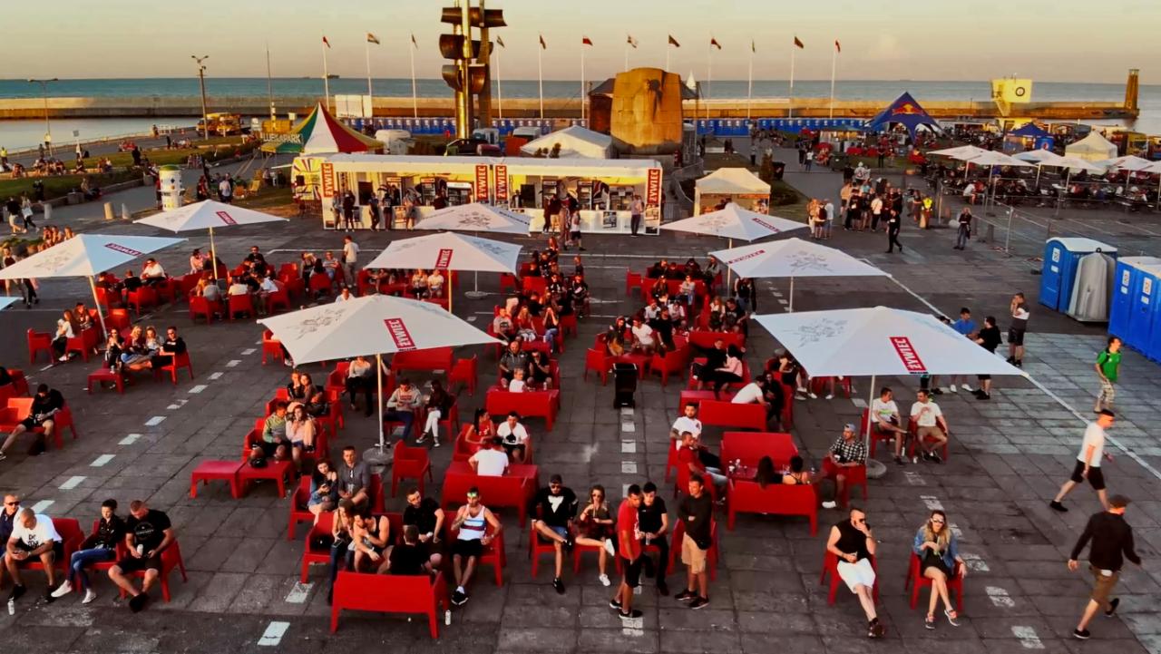 Widok z góry na plenerową strefę relaksu z czerwonymi krzesłami i stolikami pod białymi parasolami z logo, w tle budki z jedzeniem i napojami, oraz nadmorski krajobraz.