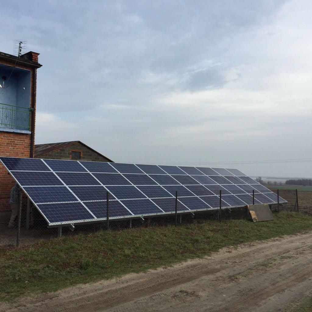 Rozległa instalacja paneli słonecznych na metalowej konstrukcji, umieszczona na polu obok ceglanego domu z balkonem, pod pochmurnym niebem.