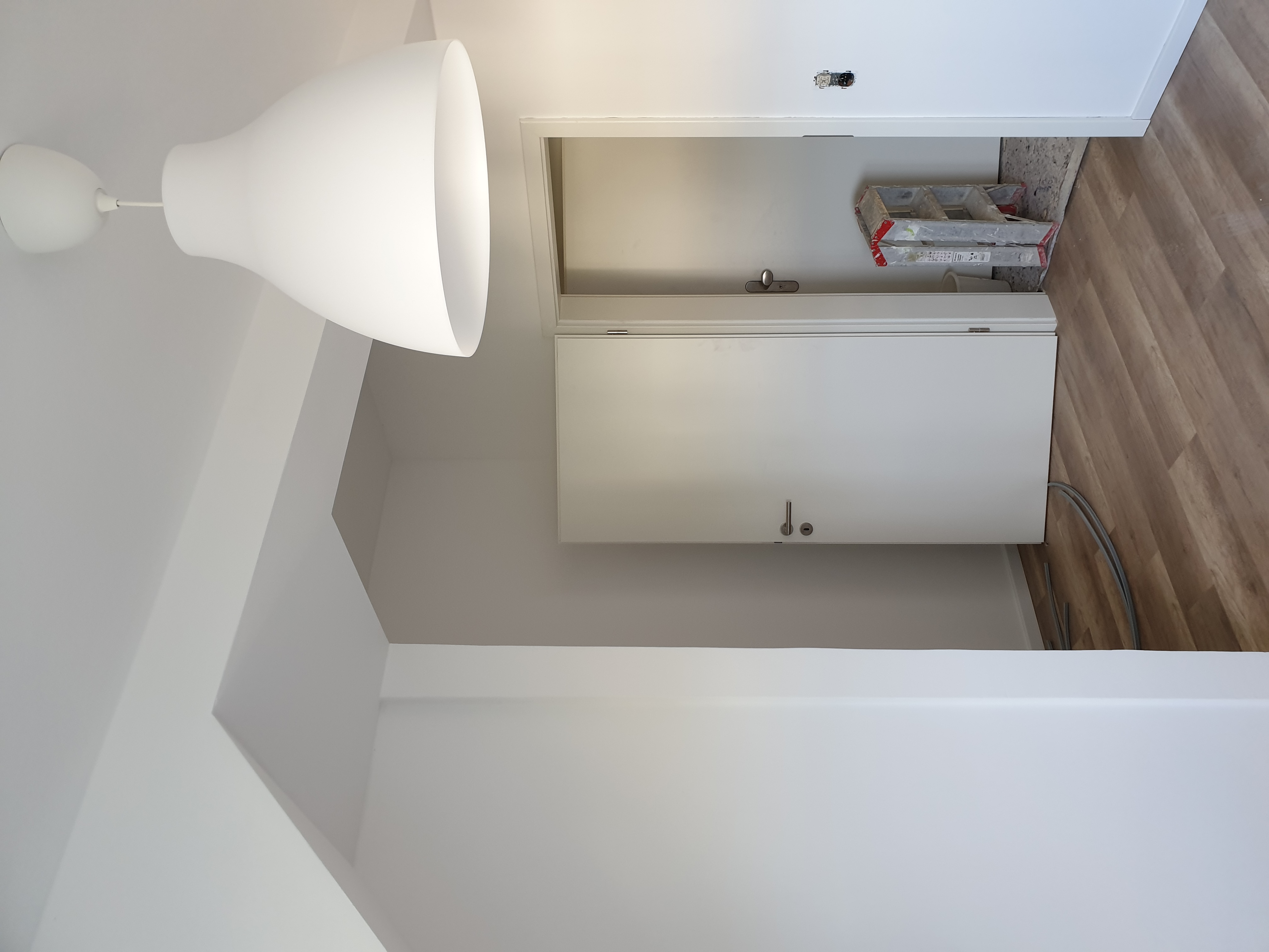 Wnętrze w trakcie odświeżania: białe ściany, drewnopodobna podłoga, wisząca lampa z tworzywa, drabina malarska w otwartym pomieszczeniu z widocznym fragmentem instalacji elektrycznej.