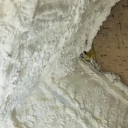 Poddasze z częściowo nałożoną białą pianką izolacyjną PUR, widoczne fragmenty ceglanej ściany i drewnianej konstrukcji dachu.