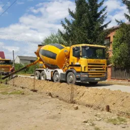 Żółta betonomieszarka Scania z czarnymi pasami na bębnie, w trakcie wylewania betonu do przygotowanego wykopu z wystającymi prętami zbrojeniowymi, w tle drugi pojazd i pracownicy.