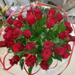 Annabelle - Bukiet czerwonych róż owinięty przezroczystą folią z czerwoną wstążką, na białym blacie, w tle fragment wystawy kwiaciarni.