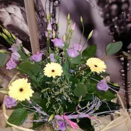 Annabelle - Kompozycja kwiatowa z jasnożółtych gerber, fioletowych eustom i zielonych liści w wiklinowym koszyku, na tle abstrakcyjnej grafiki w odcieniach szarości.