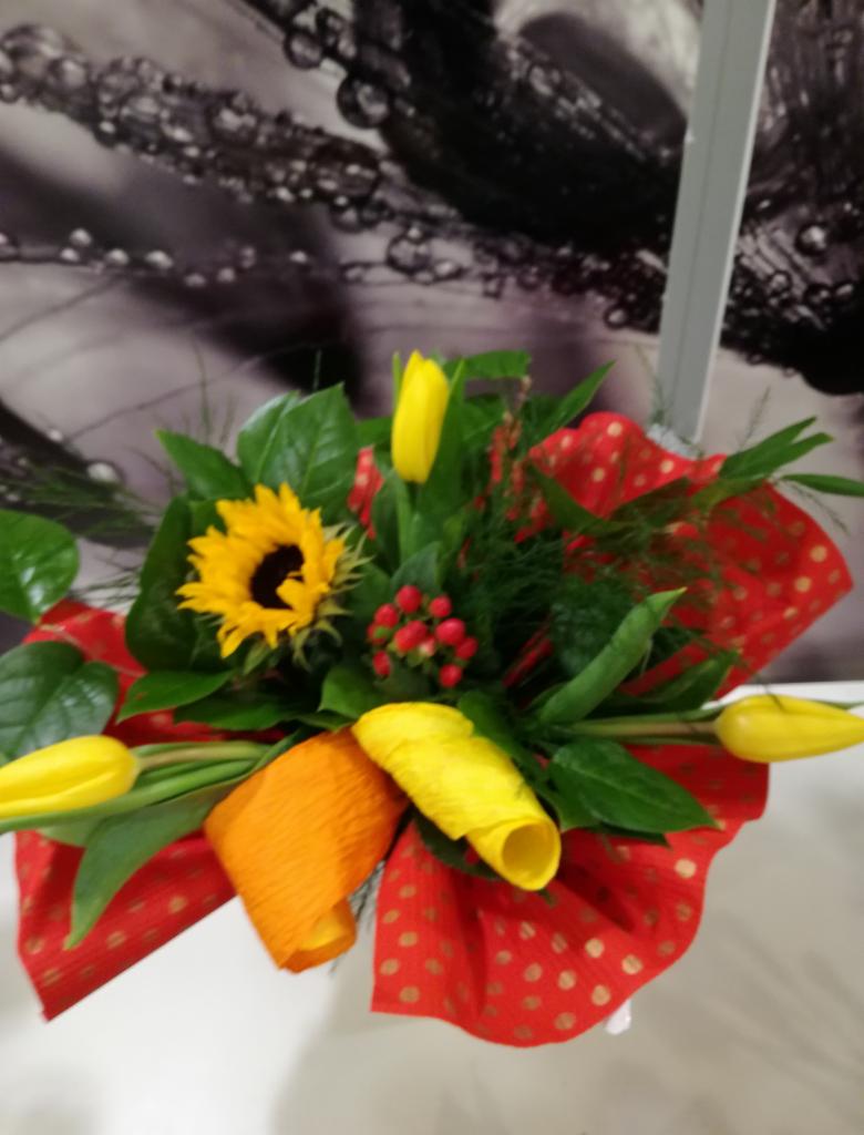 Kompozycja kwiatowa z żółtych tulipanów, słonecznika i czerwonych jagód, owinięta w czerwoną tkaninę w złote kropki, na tle rozmytej grafiki z motywem kropli.