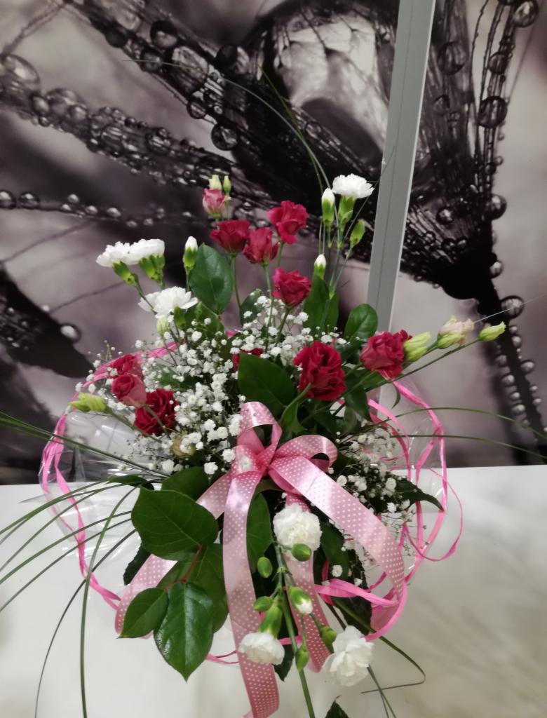 Kompozycja kwiatowa z czerwonych różyczek i białych goździków w przezroczystym owalu, ozdobiona różową wstążką w białe kropki, na tle grafiki z motywem kropli.