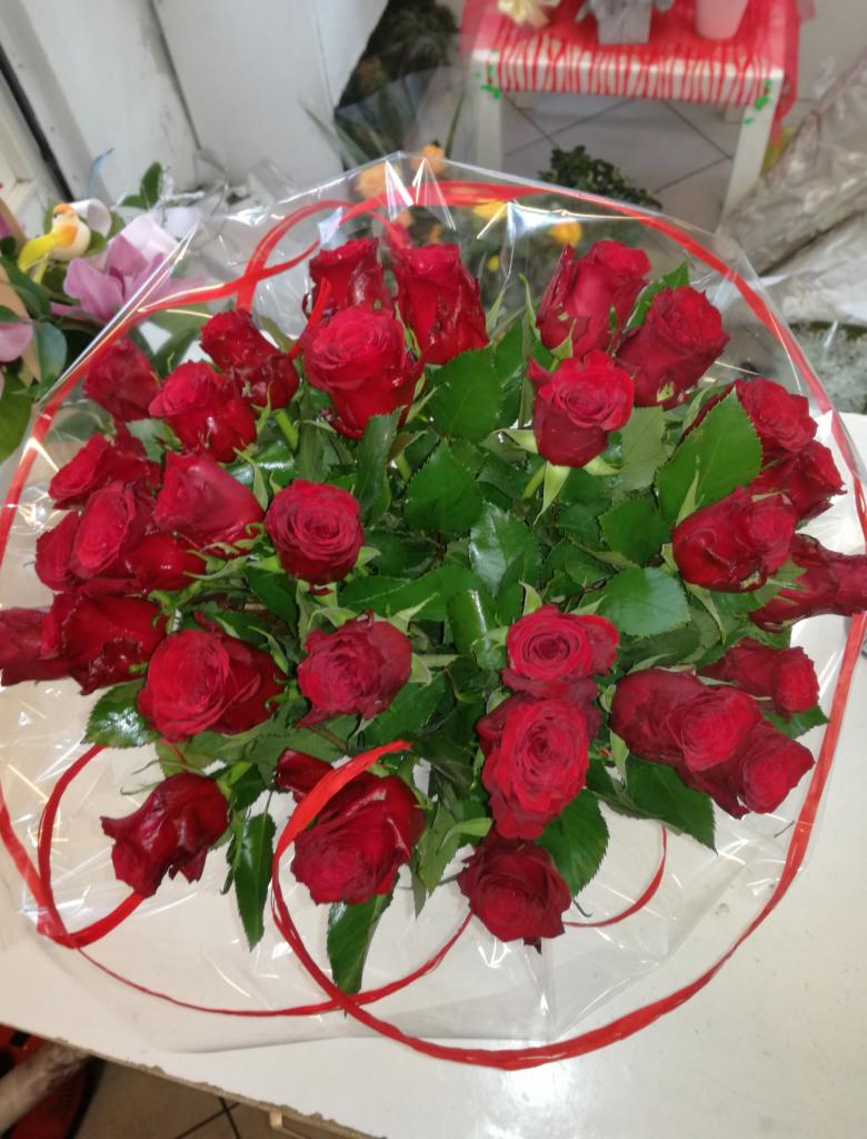 Bukiet czerwonych róż owinięty przezroczystą folią z czerwoną wstążką, na białym blacie, w tle fragment wystawy kwiaciarni.