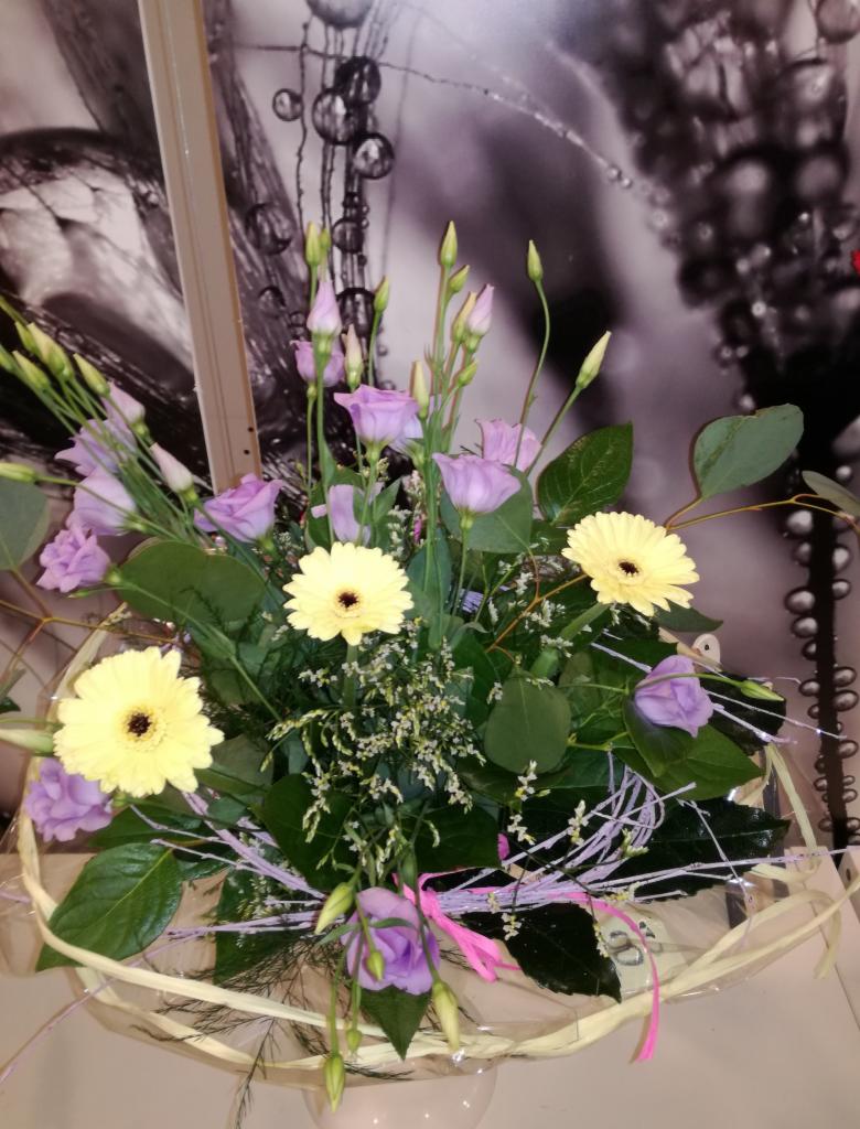 Kompozycja kwiatowa z jasnożółtych gerber, fioletowych eustom i zielonych liści w wiklinowym koszyku, na tle abstrakcyjnej grafiki w odcieniach szarości.