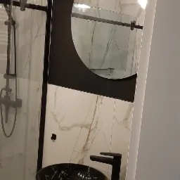 Nowoczesna łazienka z czarną umywalką nablatową w marmurkowy wzór i czarną armaturą, na blacie z jasnego marmuru. W tle kabina prysznicowa z czarnymi ramami i okrągłe lustro z czarną obudową.