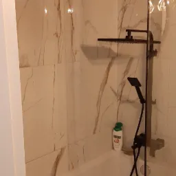 Nowoczesna kabina prysznicowa nad wanną z czarną armaturą i marmurowymi płytkami, widok na butelkę kosmetyku na półce.