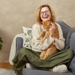 Uśmiechnięta kobieta w okularach siedzi na szarej kanapie z rudym kotem na kolanach, w tle roślina doniczkowa i beżowa ściana.