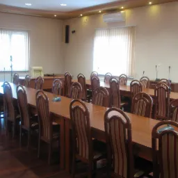 Sala konferencyjna z długimi drewnianymi stołami, krzesłami w paski i mikrofonami na stołach, oświetlona światłem dziennym z okien.