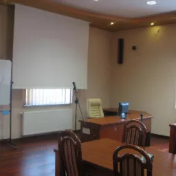 Sala szkoleniowa z drewnianymi stołami, krzesłami z czerwonymi paskami, biurkiem z komputerem, mikrofonem na statywie, tablicą suchościeralną i opuszczonym ekranem projekcyjnym.