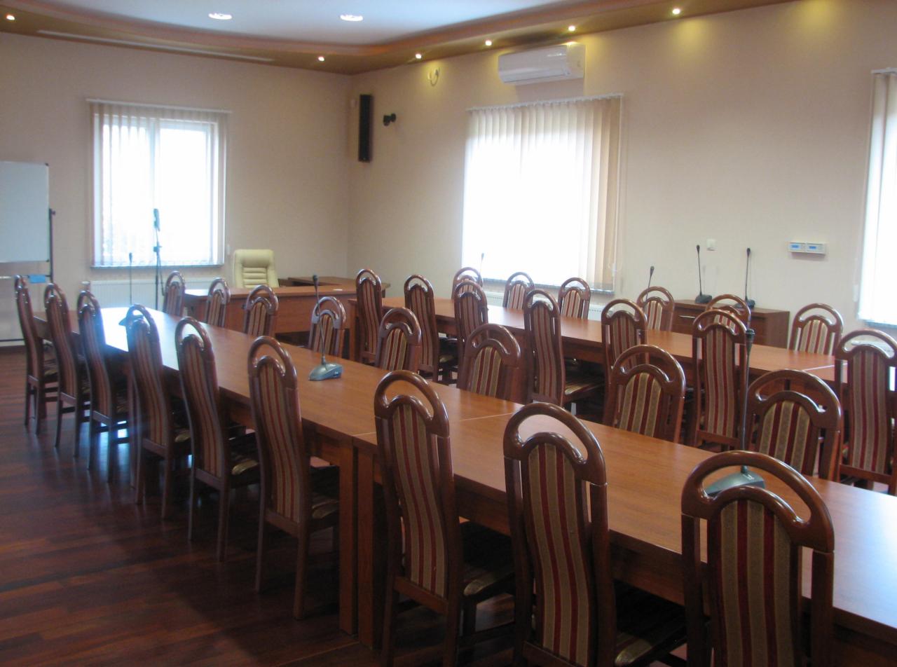 Sala konferencyjna z długimi drewnianymi stołami, krzesłami w paski i mikrofonami na stołach, oświetlona światłem dziennym z okien.