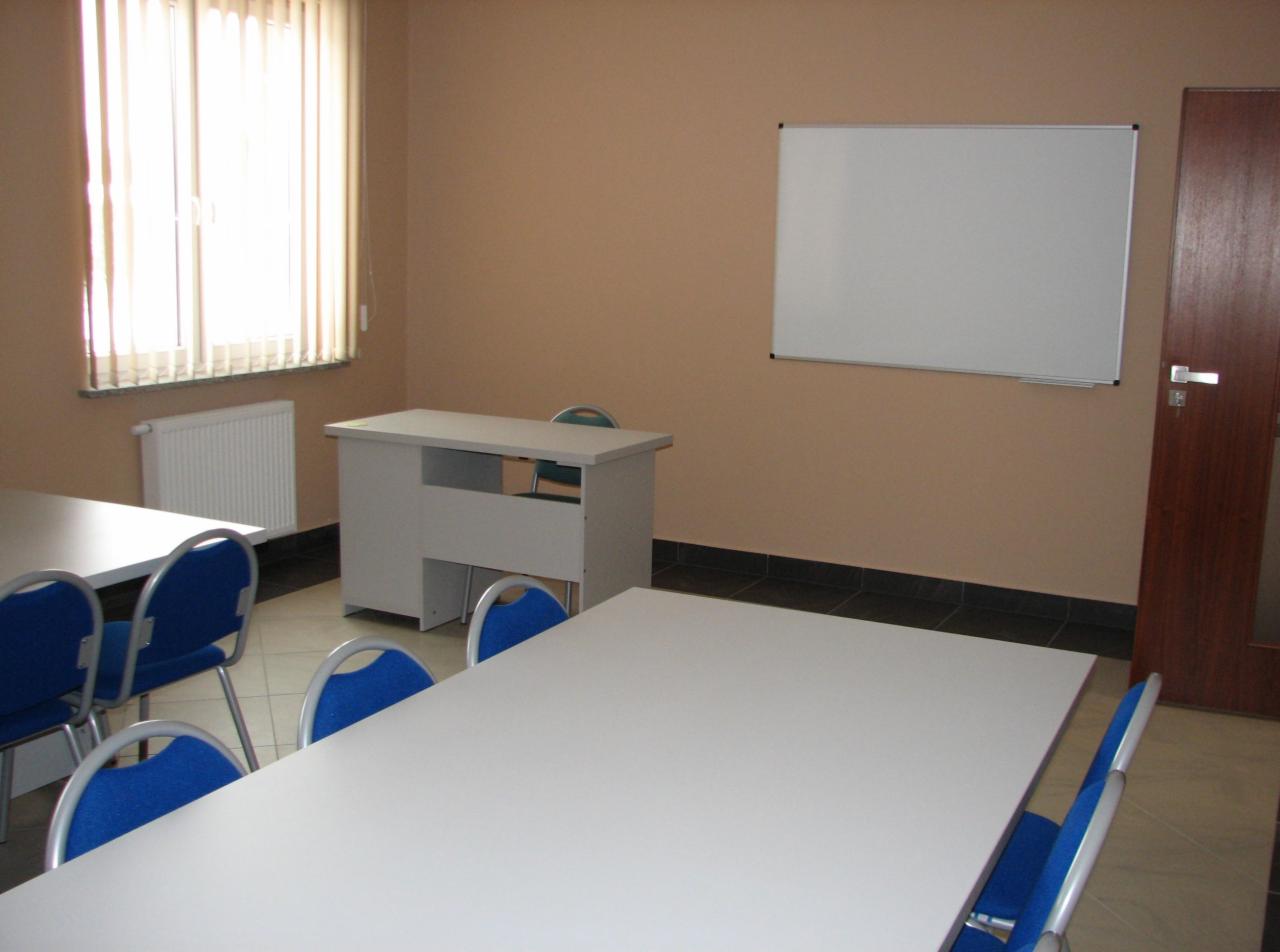 Pusta sala szkoleniowa z białymi stołami, niebieskimi krzesłami, tablicą suchościeralną i zasłoniętym oknem, przygotowana do zajęć.