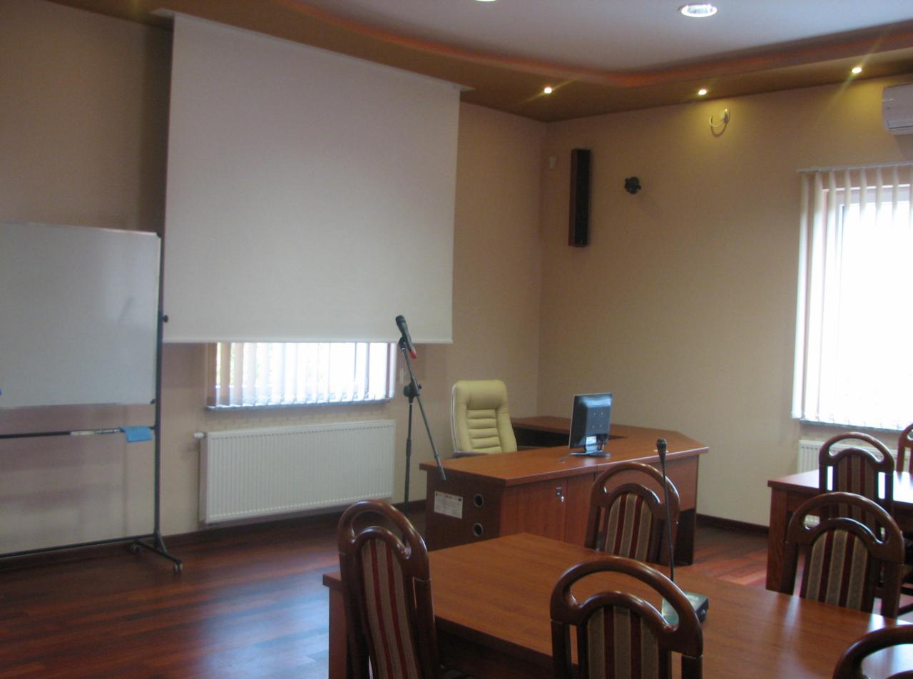 Sala szkoleniowa z drewnianymi stołami, krzesłami z czerwonymi paskami, biurkiem z komputerem, mikrofonem na statywie, tablicą suchościeralną i opuszczonym ekranem projekcyjnym.
