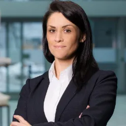 Portret kobiety w ciemnym garniturze i białej koszuli, z rękami skrzyżowanymi, na tle przeszklonego biura.