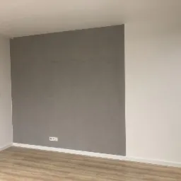 Wykończone wnętrze pokoju z szarą ścianą pokrytą gładzią dekoracyjną, białą ścianą z włącznikami i gniazdami, oraz podłogą z paneli imitujących drewno.