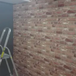 Ściana z cegieł dekoracyjnych w trakcie montażu, widoczna drabina aluminiowa i stół z narzędziami pomiarowymi, ciemne belki na suficie.