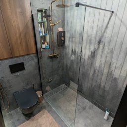 BergerDesign - Nowoczesna łazienka z kabiną prysznicową typu walk-in, szarą armaturą i płytkami z motywem lasu. Uwagę przykuwa złota deszczownica i minimalistyczna toaleta.