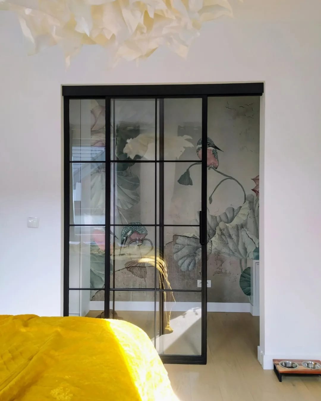Nowoczesne, przesuwne drzwi szklane z czarnymi szprosami, oddzielające pokój z żółtym pledem od wnętrza z tapetą w motywy roślinne i ptasie. Minimalistyczny design.