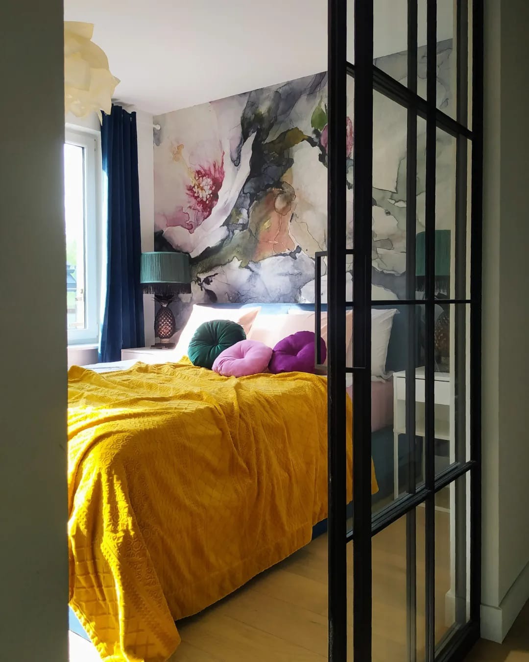 Sypialnia z żółtą narzutą, okrągłymi poduszkami i ścianą z motywem kwiatowym, widziana przez przeszkloną ściankę działową w stylu industrialnym.